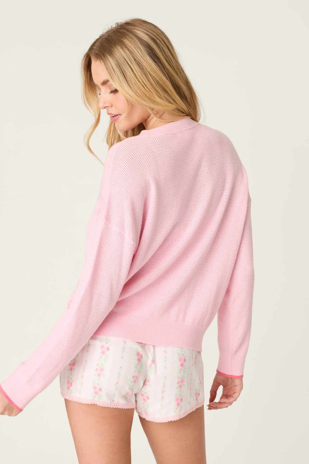 A woman with long blonde hair wears the P.J. Salvage Roses Are Pink Rosé All Day Snuggle Sweater, paired with white floral shorts. She looks over her shoulder, turned slightly away against a plain light background.