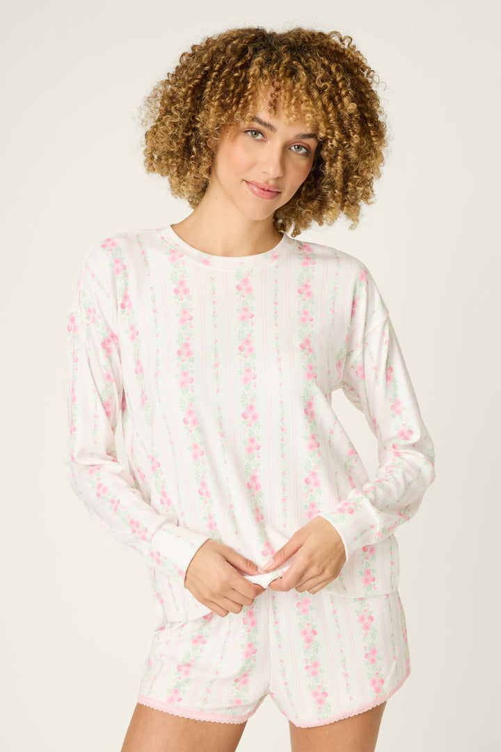 A person with curly hair models the P.J. Salvage Roses Are Pink Petal Dream Pointelle Long Sleeve Top, featuring vertical pink rose floral stripes on white, against a plain light background.