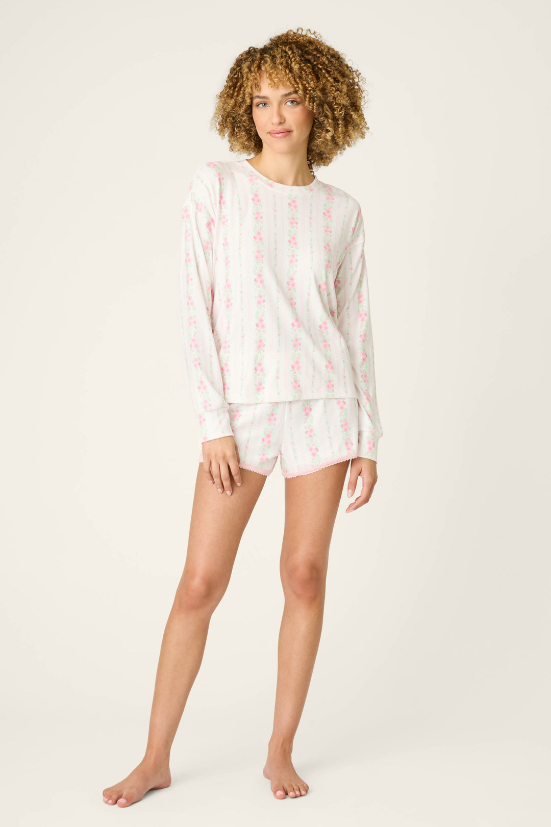 A person stands barefoot on a light background, wearing the Roses Are Pink Petal Dream Pointelle Long Sleeve Top by P.J. Salvage and matching shorts in a delicate rose floral print.