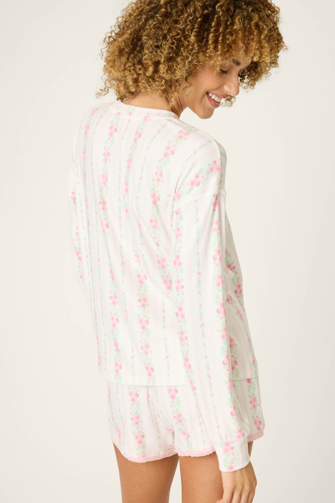 A person with curly hair smiles over their shoulder while wearing the P.J. Salvage Roses Are Pink Petal Dream Pointelle Long Sleeve Top, featuring a rose floral print. They stand against a plain, light background.