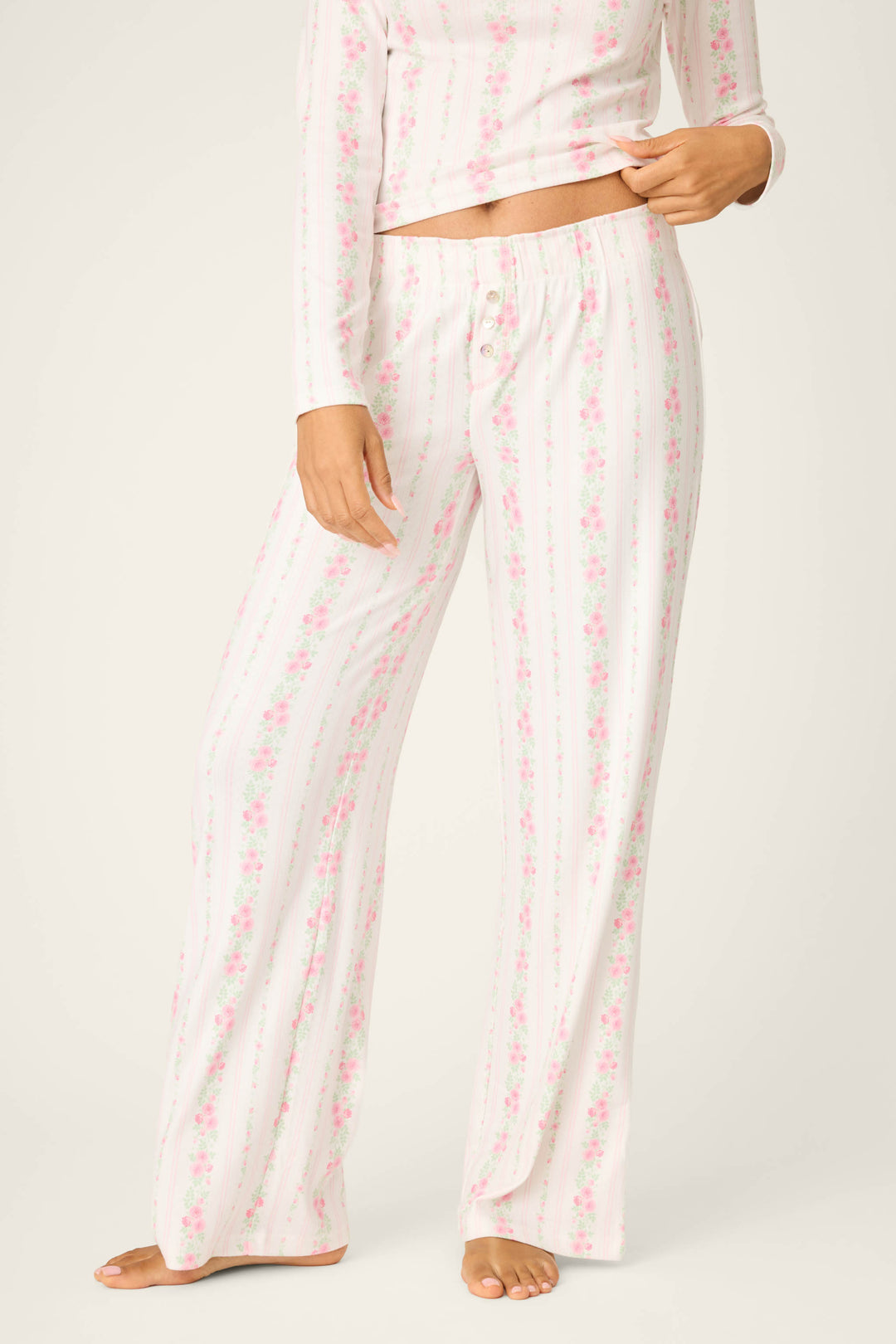 A person wears P.J. Salvage Roses Are Pink Petal Dream Pointelle Open Leg Pants—white with vertical pink stripes and a floral print—crafted from soft double brushed pointelle. The wearer is barefoot, shown only from the waist down.