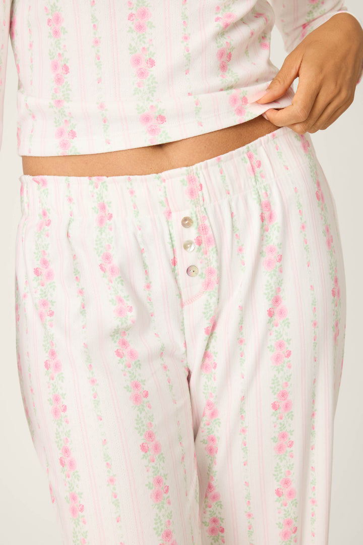 A person wearing P.J. Salvage Roses Are Pink Petal Dream Pointelle Open Leg Pants, featuring a floral print and three-button waistband, lifts the pajama top hem slightly with one hand.