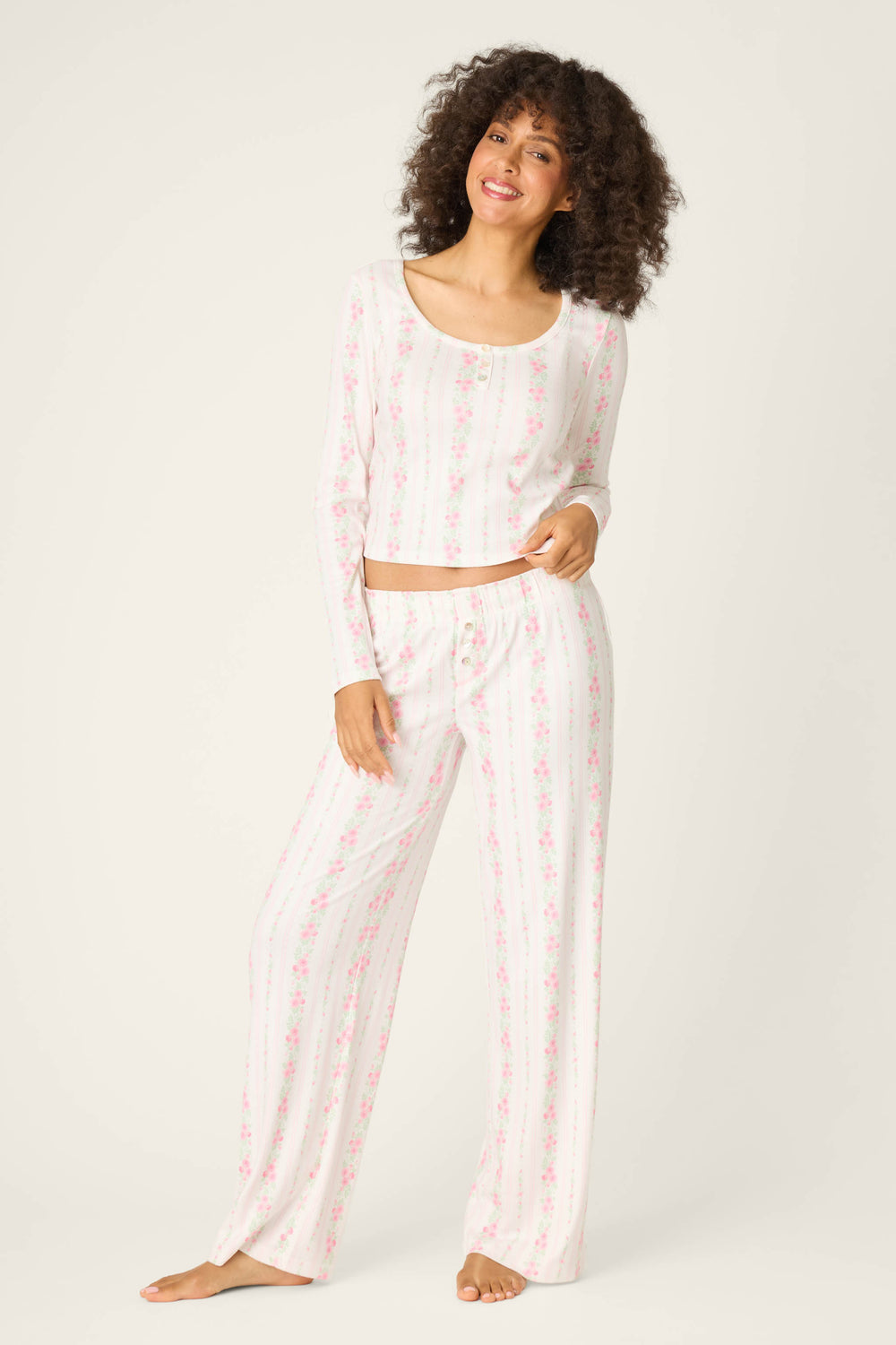 A woman stands barefoot against a light background, smiling with one hand on her hip, wearing P.J. Salvage Roses Are Pink Petal Dream Pointelle Open Leg Pants and matching long-sleeve top with a pink and white floral striped pattern.