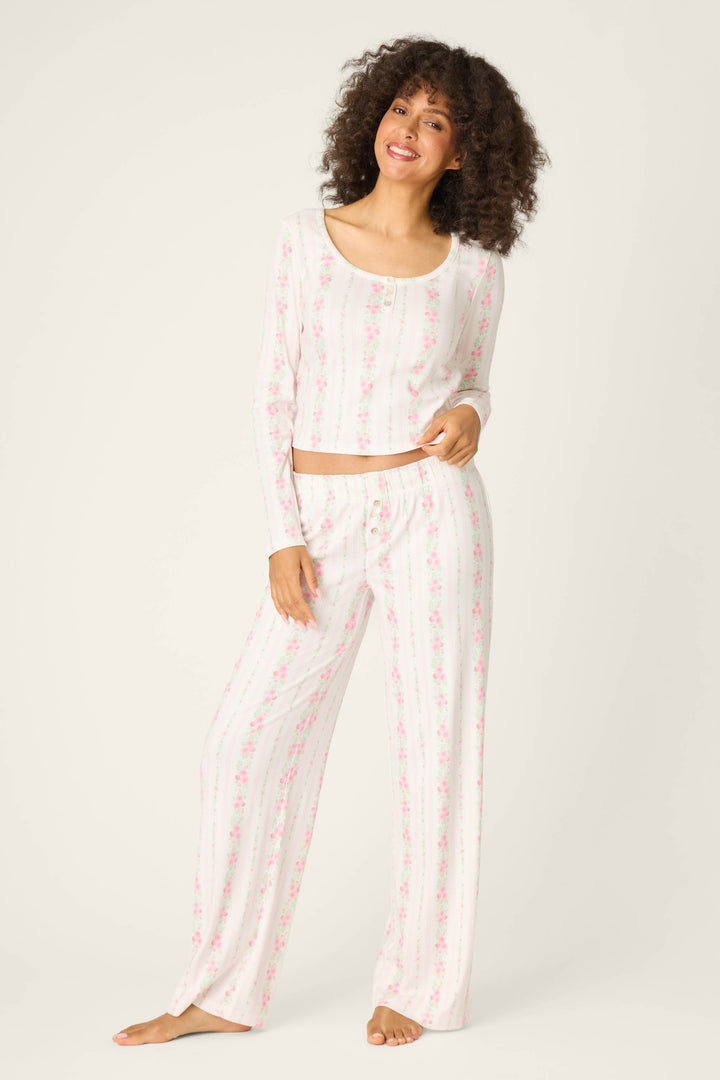 A woman stands barefoot against a light background, smiling with one hand on her hip, wearing P.J. Salvage Roses Are Pink Petal Dream Pointelle Open Leg Pants and matching long-sleeve top with a pink and white floral striped pattern.