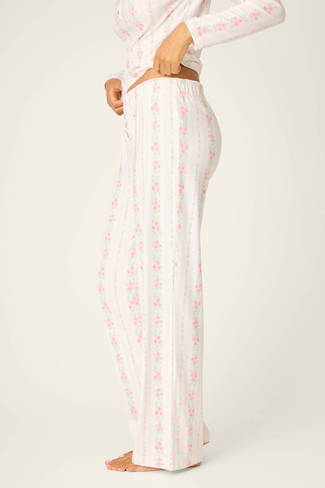 A person stands barefoot in a side view against a plain background, wearing P.J. Salvage Roses Are Pink Petal Dream Pointelle Open Leg Pants with a matching floral long-sleeve top.