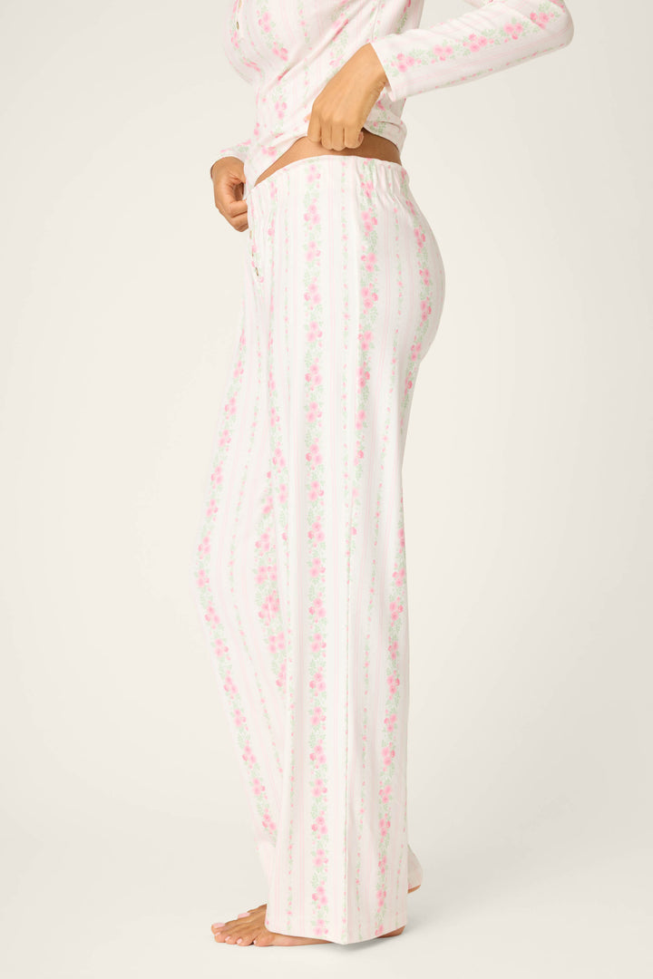 A person stands barefoot in a side view against a plain background, wearing P.J. Salvage Roses Are Pink Petal Dream Pointelle Open Leg Pants with a matching floral long-sleeve top.