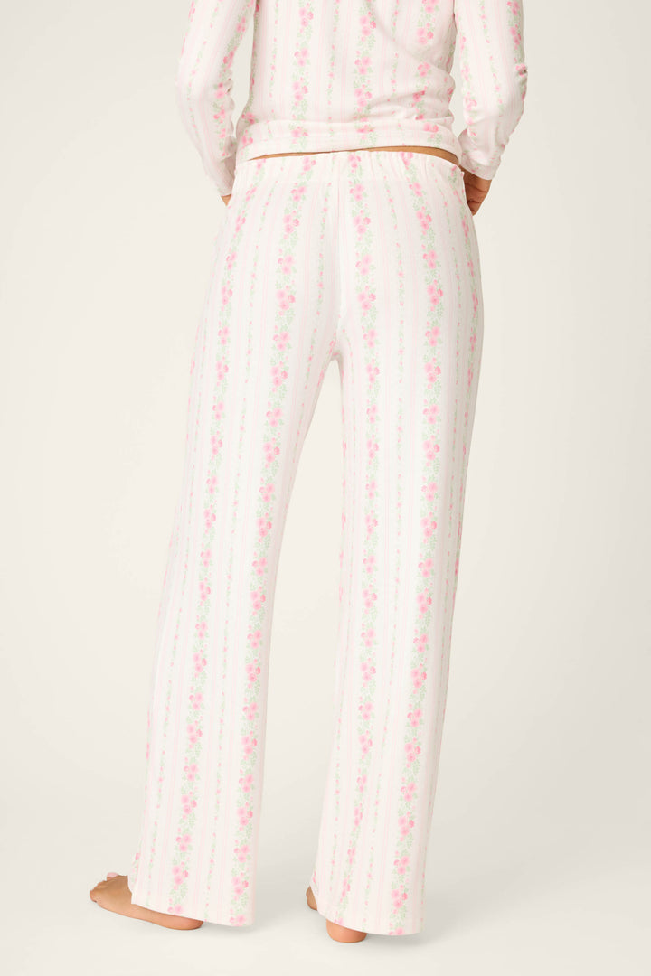 A person stands barefoot, facing away from the camera in P.J. Salvage Roses Are Pink Petal Dream Pointelle Open Leg Pants—light-colored with a vertical pink and green floral print, crafted from soft double brushed pointelle. The background is plain and neutral.