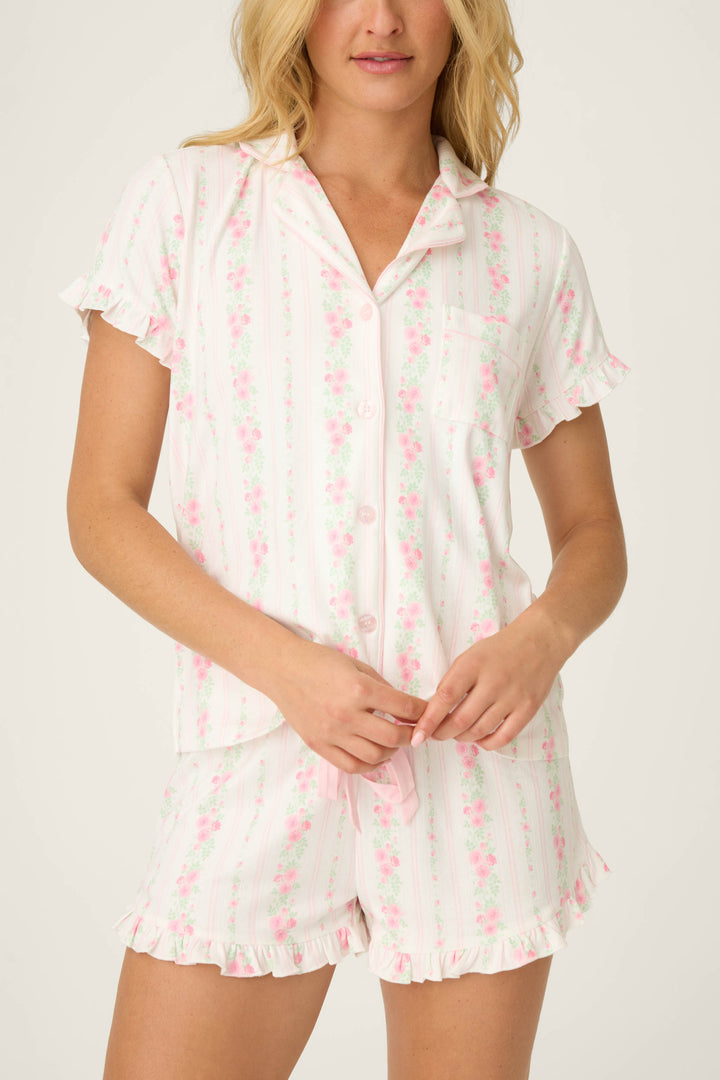 A person models the P.J. Salvage Roses Are Pink Petal Dream Pointelle 3-Piece PJ Set, featuring a short-sleeved, button-up top and matching pink floral shorts with ruffled edges. Their face is partially out of frame against a plain background.