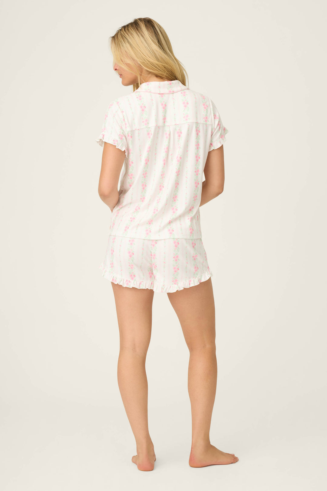 A woman stands barefoot, facing away, wearing the Roses Are Pink Petal Dream Pointelle 3-Piece PJ Set by P.J. Salvage: a white short-sleeve top and shorts with pink floral print, on a plain light background.