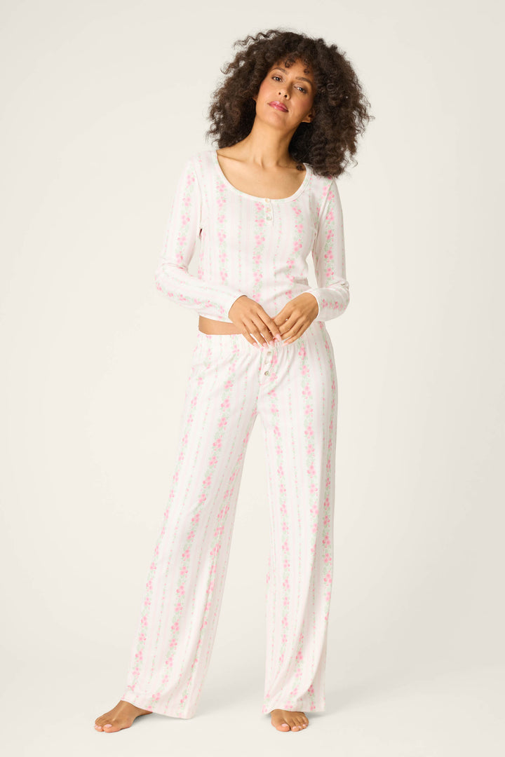 A woman with curly hair stands barefoot against a plain background, wearing the P.J. Salvage Roses Are Pink Petal Dream Pointelle PJ Set featuring a light pink and white floral print, looking relaxed.