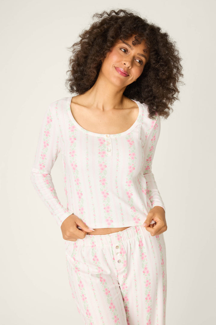 A person with curly hair smiles gently while wearing the P.J. Salvage Roses Are Pink Petal Dream Pointelle PJ Set, a long-sleeved white floral pajama with pink patterns, and holds the shirt’s hem against a plain background.