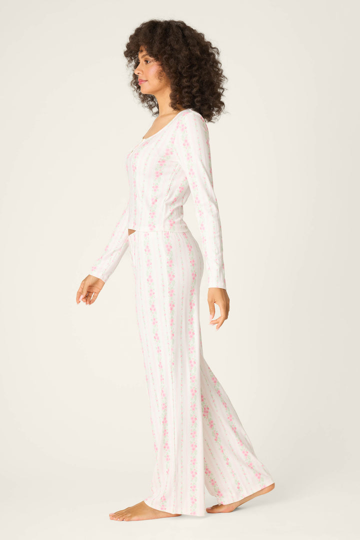 A woman stands in profile wearing the P.J. Salvage Roses Are Pink Petal Dream Pointelle PJ Set—a long-sleeved, white pajama set with pink floral stripes and a fitted top and pants—barefoot against a plain, light background.