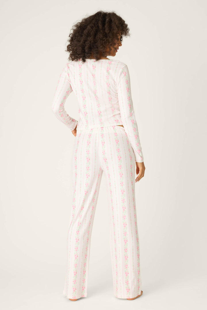 A person faces away, wearing the P.J. Salvage Roses Are Pink Petal Dream Pointelle PJ Set—a lightweight floral pajama set with vertical pink stripes on a white background, featuring a long-sleeve top and loose pants against a plain backdrop.