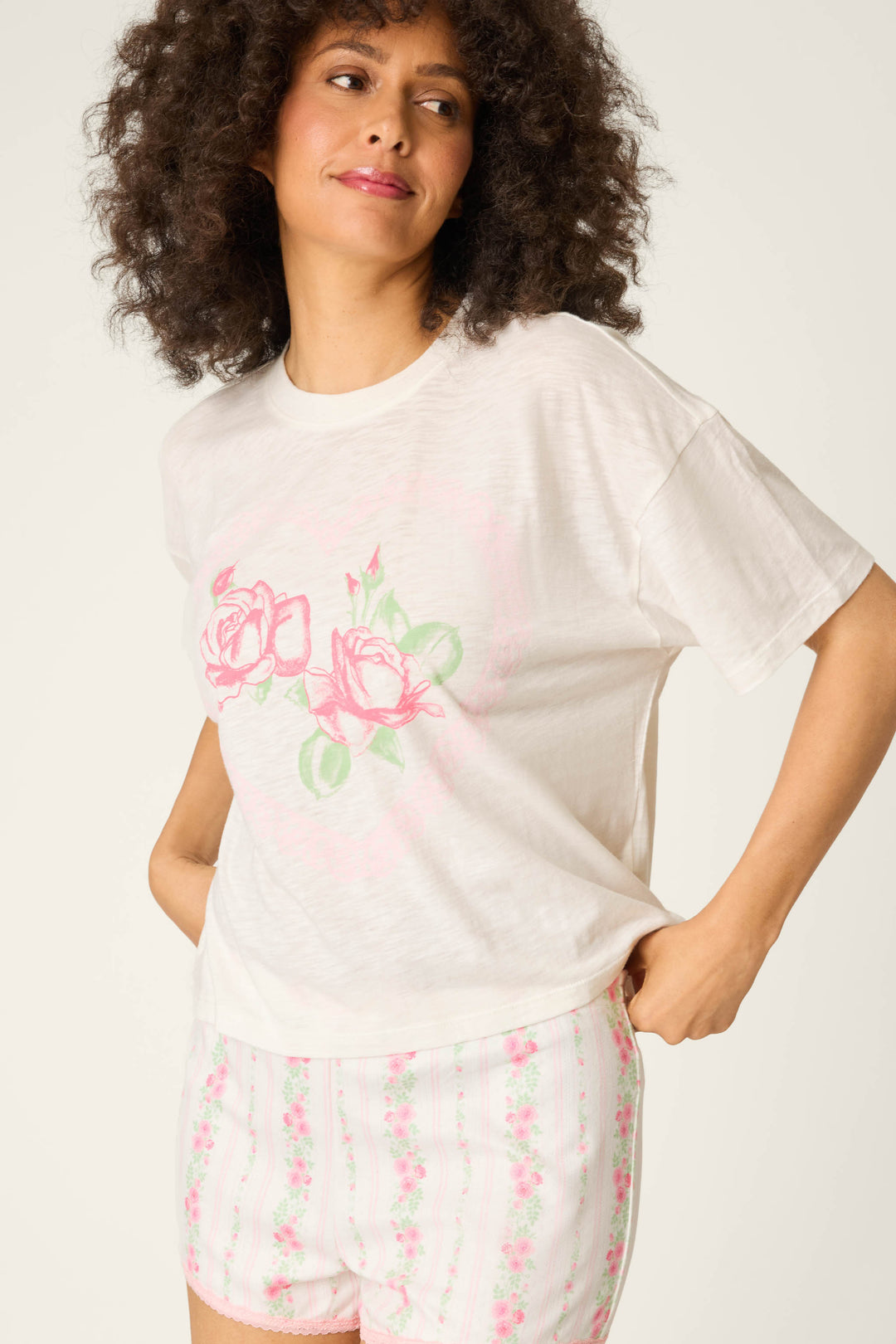 A person with curly hair wears the P.J. Salvage Roses Are Pink Heart Bloom Graphic T-Shirt and matching floral shorts, posing with one hand on their hip against a plain light background.