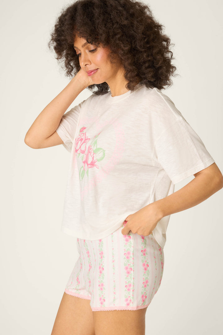 A person with curly hair wears the P.J. Salvage Roses Are Pink Heart Bloom Graphic T-Shirt and matching white shorts with pink and green floral stripes, standing sideways with one hand touching their hair.