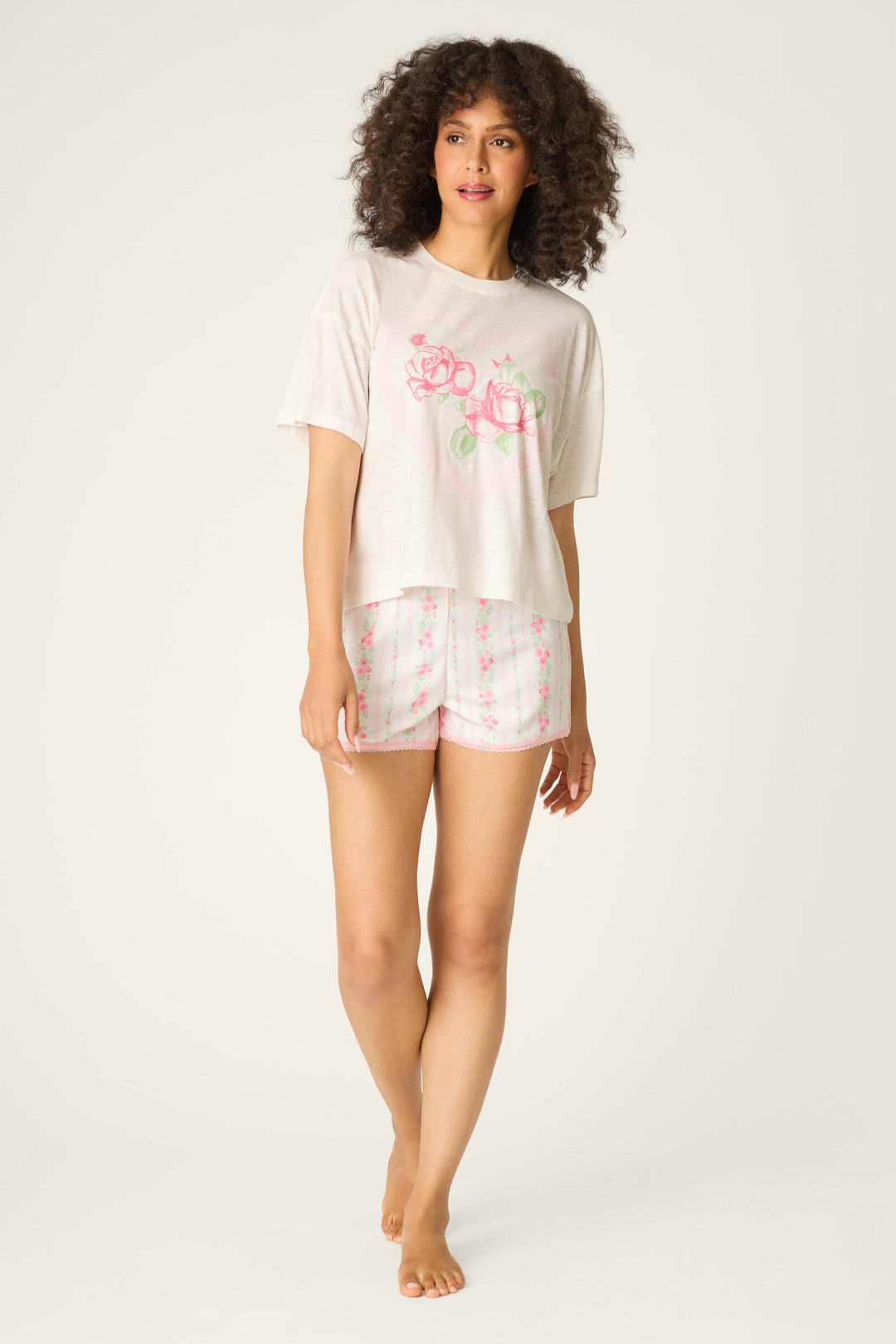 A person with curly hair stands barefoot on a light background, wearing the P.J. Salvage Roses Are Pink Heart Bloom Graphic T-Shirt and matching white floral shorts.
