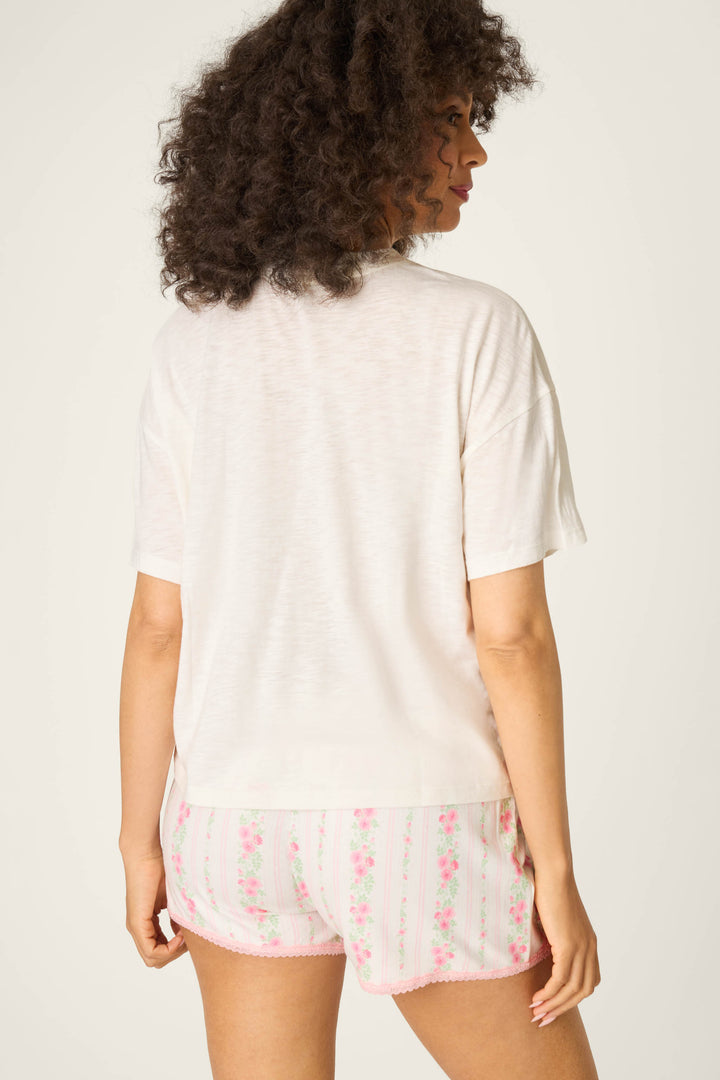 A person with curly hair stands facing away from the camera, wearing the P.J. Salvage Roses Are Pink Heart Bloom Graphic T-Shirt and white shorts with a pink and green floral pattern, against a plain light background.