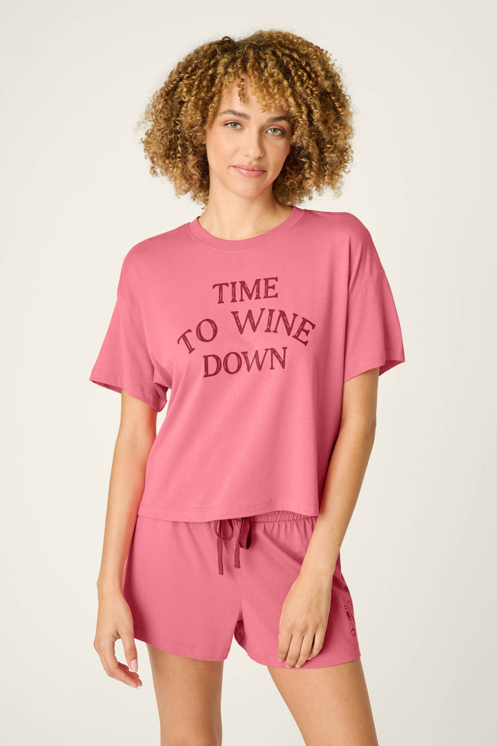 A person with curly hair models the P.J. Salvage Time to Wine Down Graphic Print Softee Set—pink pajamas featuring “TIME TO WINE DOWN” on the shirt and drawstring-waist shorts—against a plain light background.