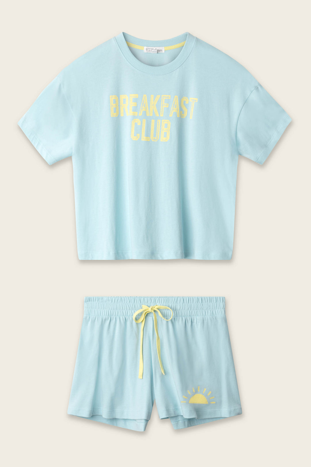 The P.J. Salvage Breakfast Club Graphic Print Softee Set features a light blue cropped T-shirt with yellow "BREAKFAST CLUB" text and matching shorts with a yellow drawstring and sunrise graphic, both laid flat on a beige background.