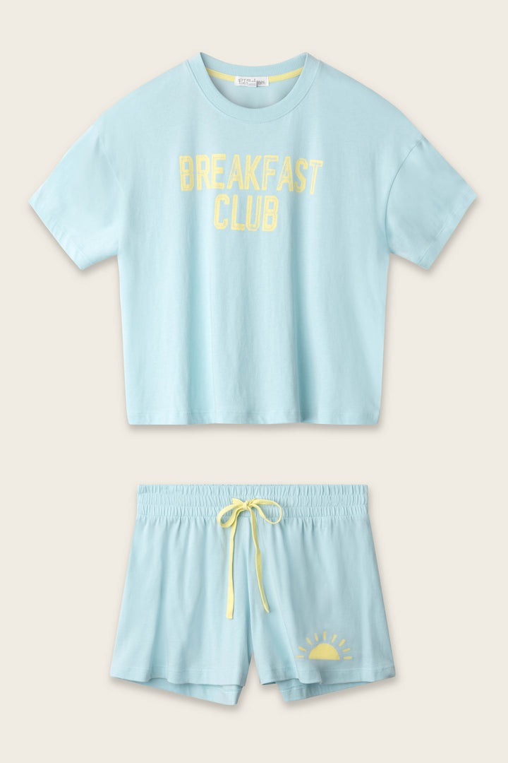 The P.J. Salvage Breakfast Club Graphic Print Softee Set features a light blue cropped T-shirt with yellow "BREAKFAST CLUB" text and matching shorts with a yellow drawstring and sunrise graphic, both laid flat on a beige background.