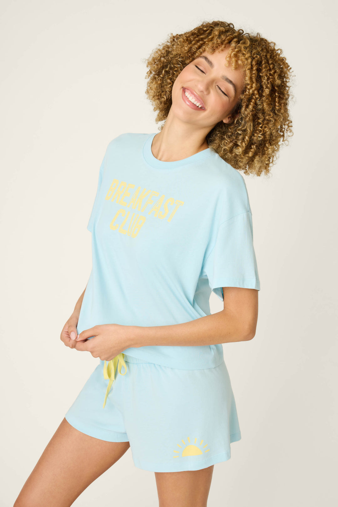 A person with curly hair smiles with eyes closed, wearing the P.J. Salvage Breakfast Club Graphic Print Softee Set—a light blue tee with "BREAKFAST CLUB" in yellow and matching shorts featuring a yellow sun graphic on one leg.