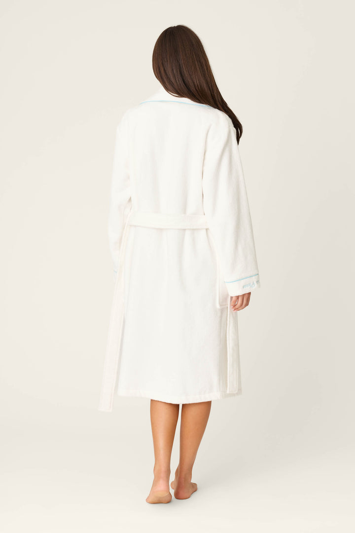 A woman with long brown hair stands barefoot, back to camera, in a knee-length white Playful Terry Happy Hour Robe by P.J. Salvage, belted at the waist against a plain light background.