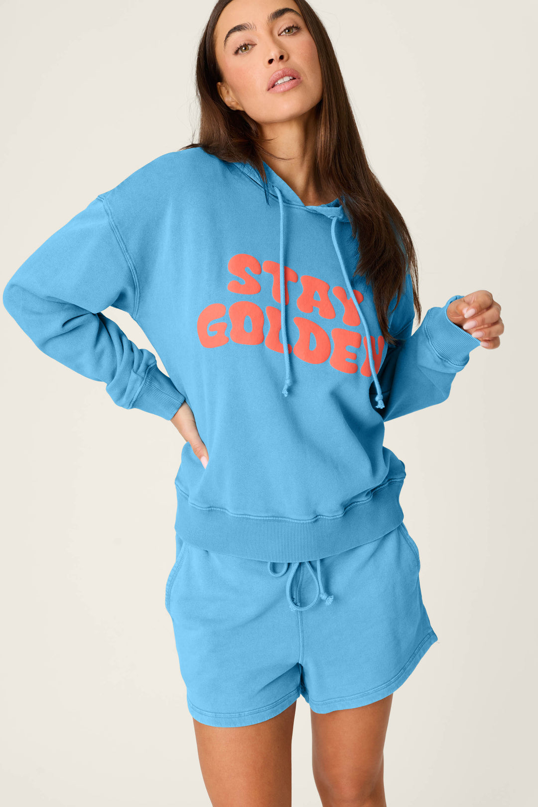 A woman models the P.J. Salvage Washed Away Stay Golden Hoodie in light blue with bold orange lettering, paired with matching shorts, standing confidently against a plain background.