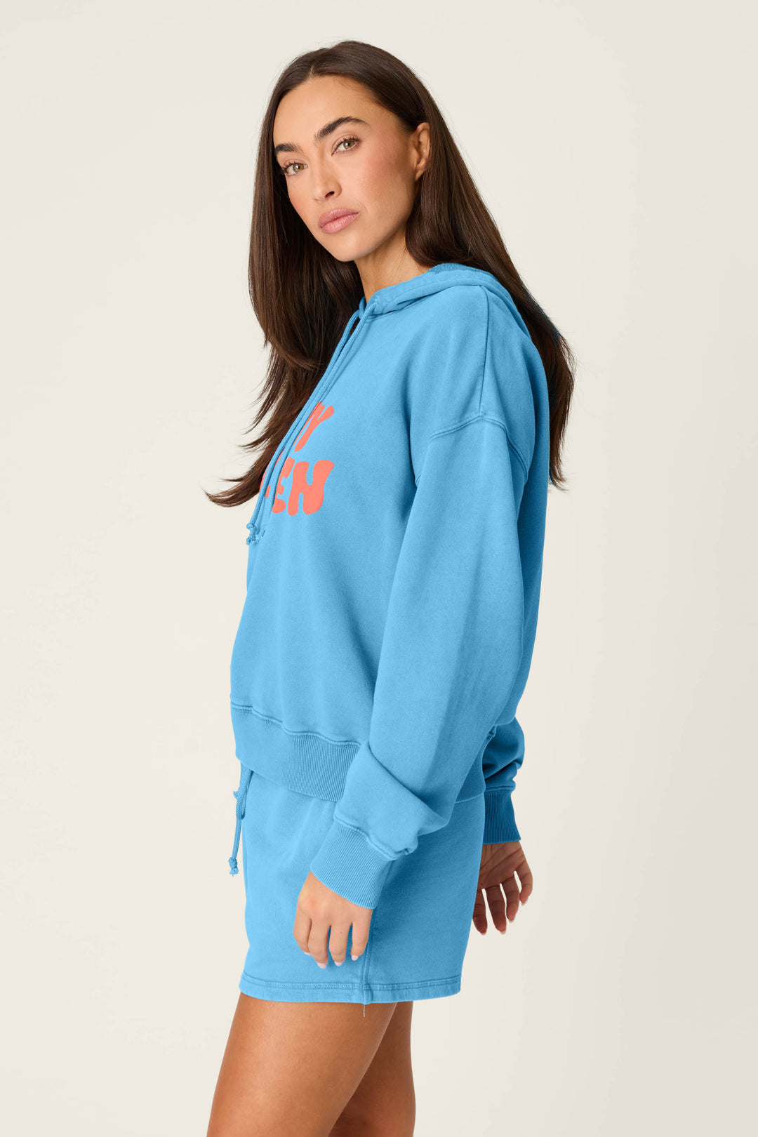 A woman with long brown hair wears the Washed Away Stay Golden Hoodie by P.J. Salvage and matching blue shorts, standing against a plain light background with a neutral expression, gazing slightly to the side.