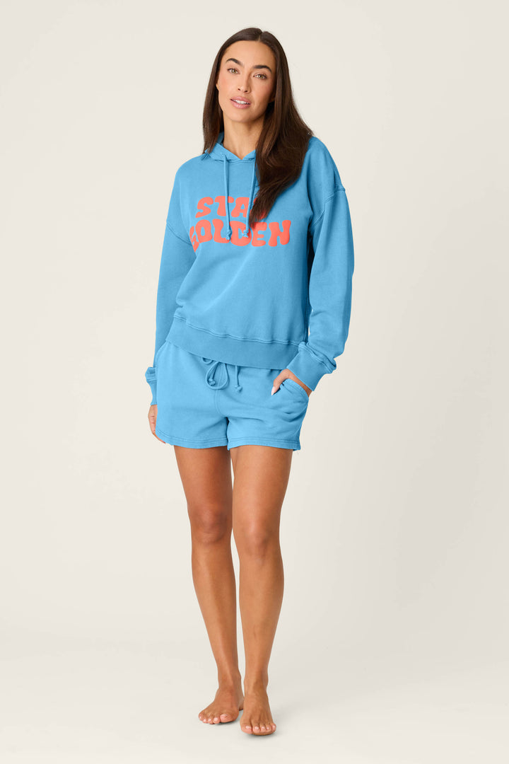 A woman with long brown hair stands barefoot against a plain background, wearing the P.J. Salvage Washed Away Stay Golden Hoodie in light blue with “Stay Golden” in orange and matching blue shorts, with one hand in her pocket.