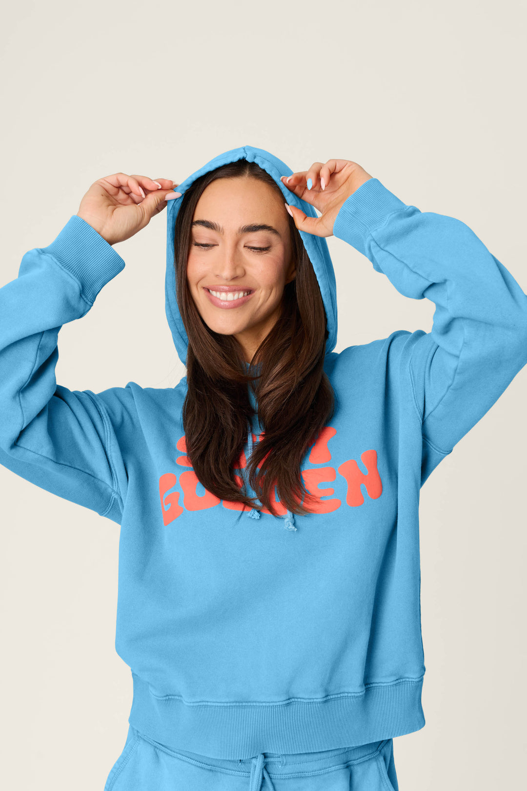 A person with long brown hair smiles with eyes closed, wearing the P.J. Salvage Washed Away Stay Golden Hoodie in bright blue with orange lettering, holding the hood with both hands against a plain light background.
