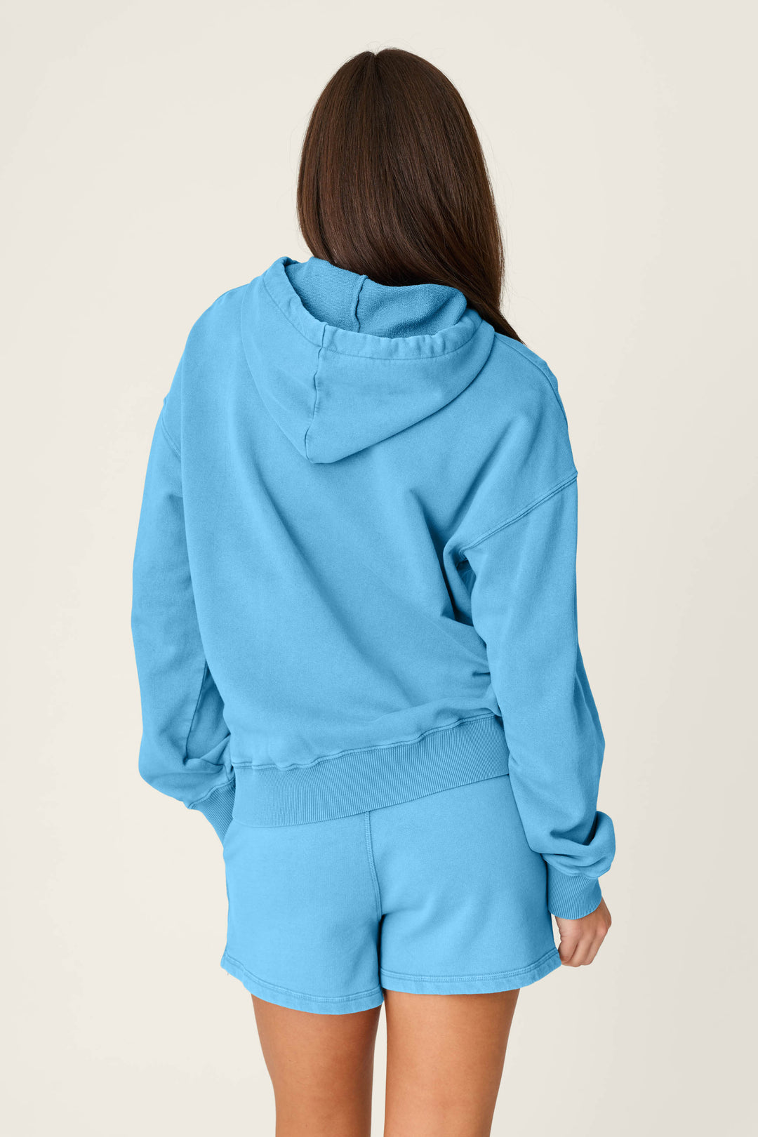 A person with long brown hair, seen from behind, is wearing the P.J. Salvage Washed Away Stay Golden Hoodie in light blue with matching shorts against a plain light background.