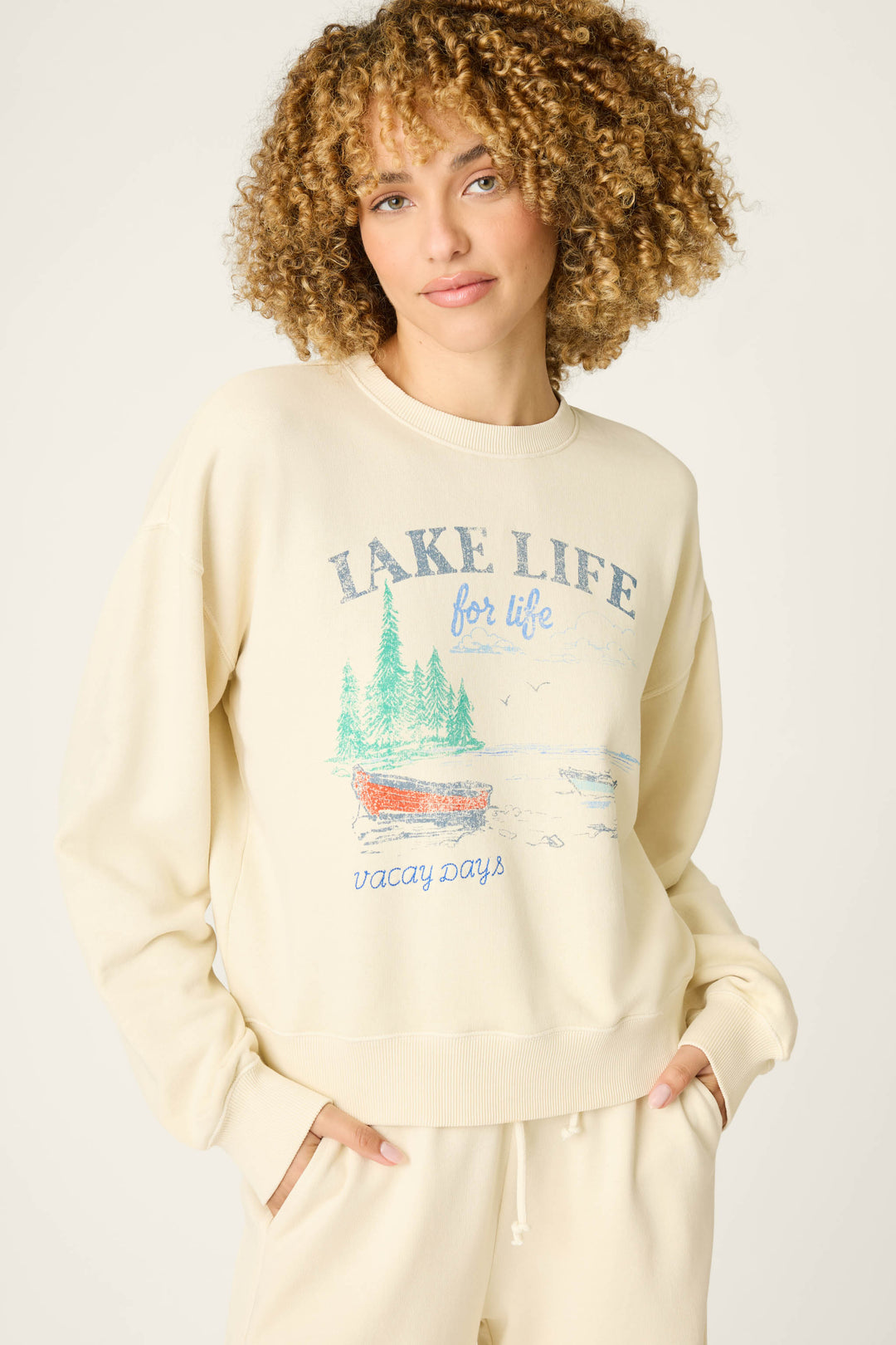A person with curly hair wears the P.J. Salvage Washed Away Lake Life For Life Pullover Sweater, cream-colored with a canoe, trees, water, and birds graphic and a relaxed fit, paired with matching pants featuring side pockets.
