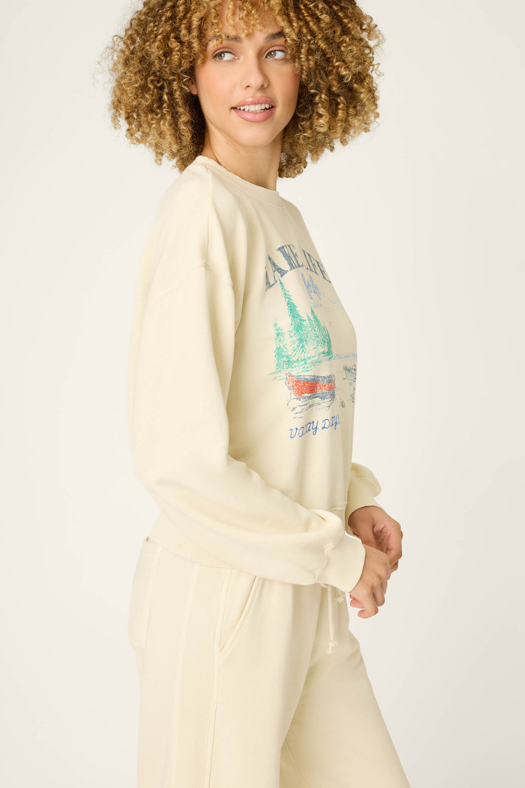 A person with curly hair wears the P.J. Salvage Washed Away Lake Life For Life Pullover Sweater, which features a canoe and trees graphic with "LAKE LIFE" and "Stay Dry" text. The background is plain and light.