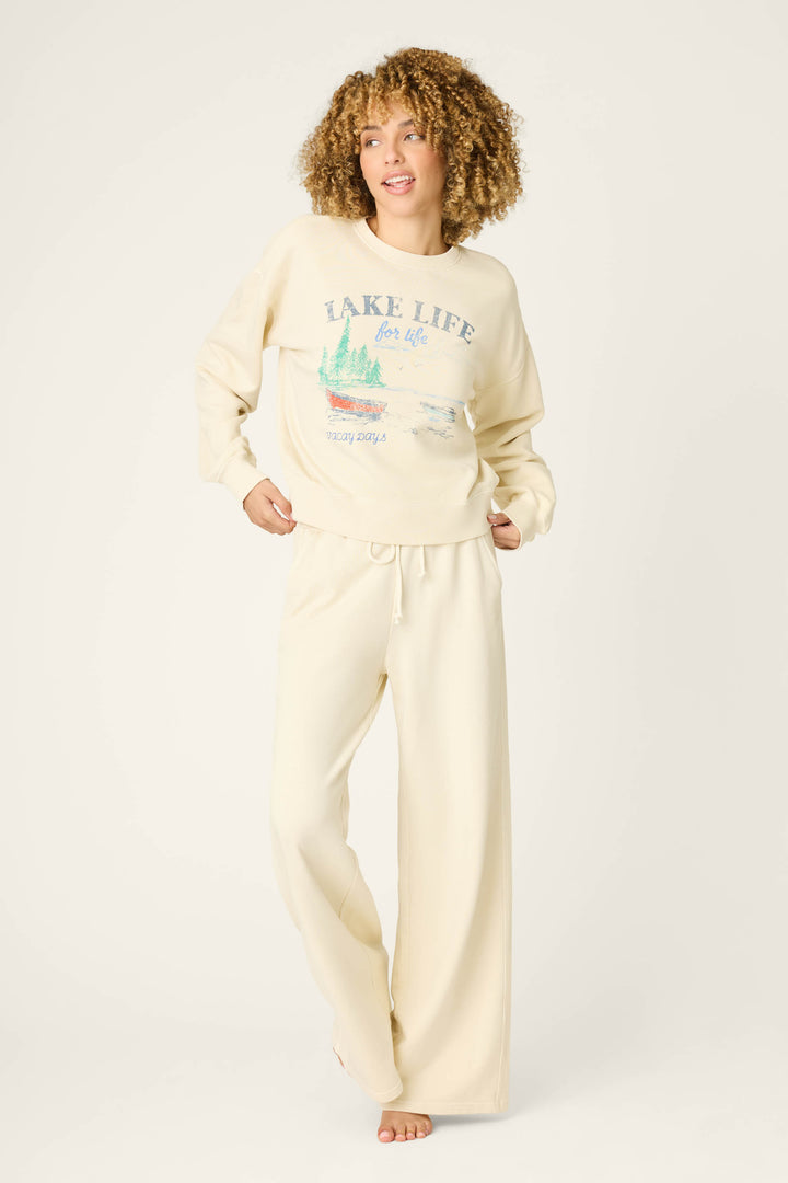 A person with curly hair models the P.J. Salvage Washed Away Lake Life For Life Pullover Sweater, cream-colored with “Lake Life for Life” and a lake-and-trees graphic, paired with matching relaxed drawstring pants, barefoot against a plain backdrop.