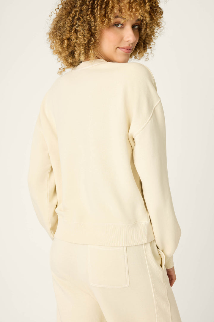 A person with curly hair is shown from the back, wearing the P.J. Salvage Washed Away Lake Life For Life Pullover Sweater, looking over their shoulder against a plain, light background.