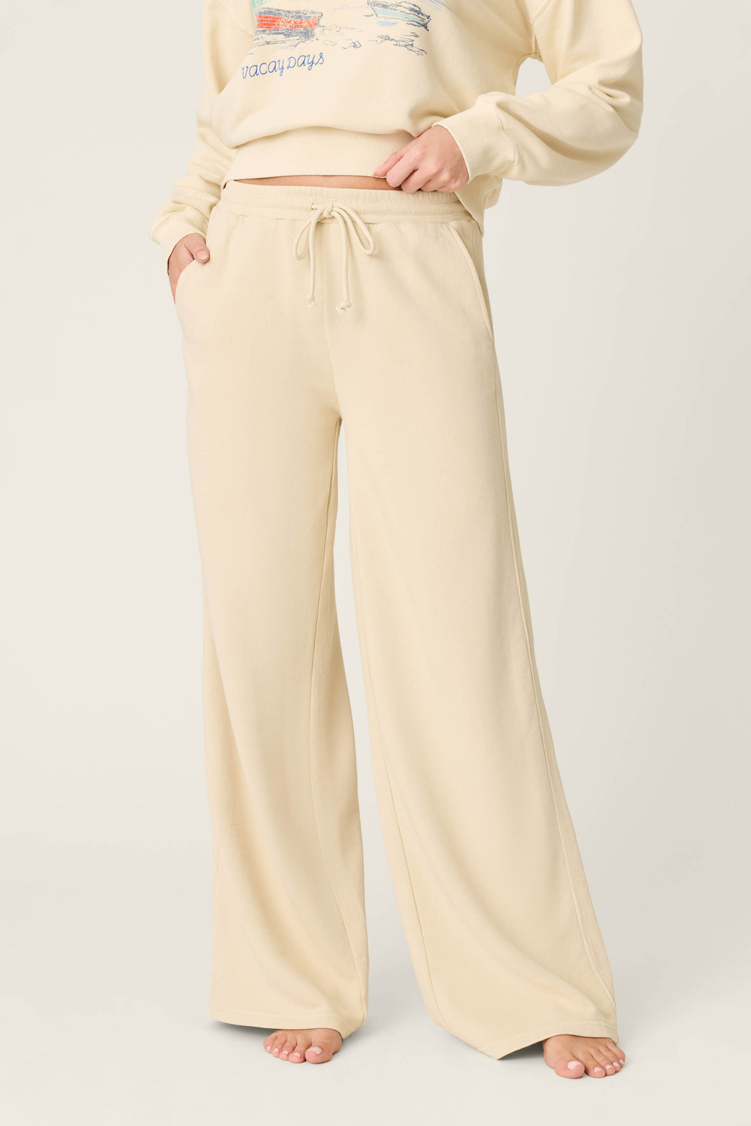 A person wears P.J. Salvage’s Washed Away Lake Life For Life Open Leg Pants, featuring a wide-leg, beige French terry design and drawstring waist, hands in pockets, barefoot against a plain light background.