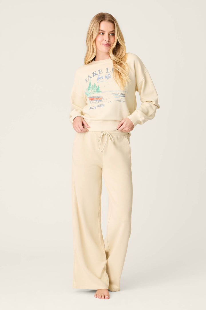 A woman with long blonde hair stands barefoot against a light background, wearing P.J. Salvage's Washed Away Lake Life For Life Open Leg Pants and a matching cream French terry sweatshirt with a graphic in a subtle sun-faded finish.