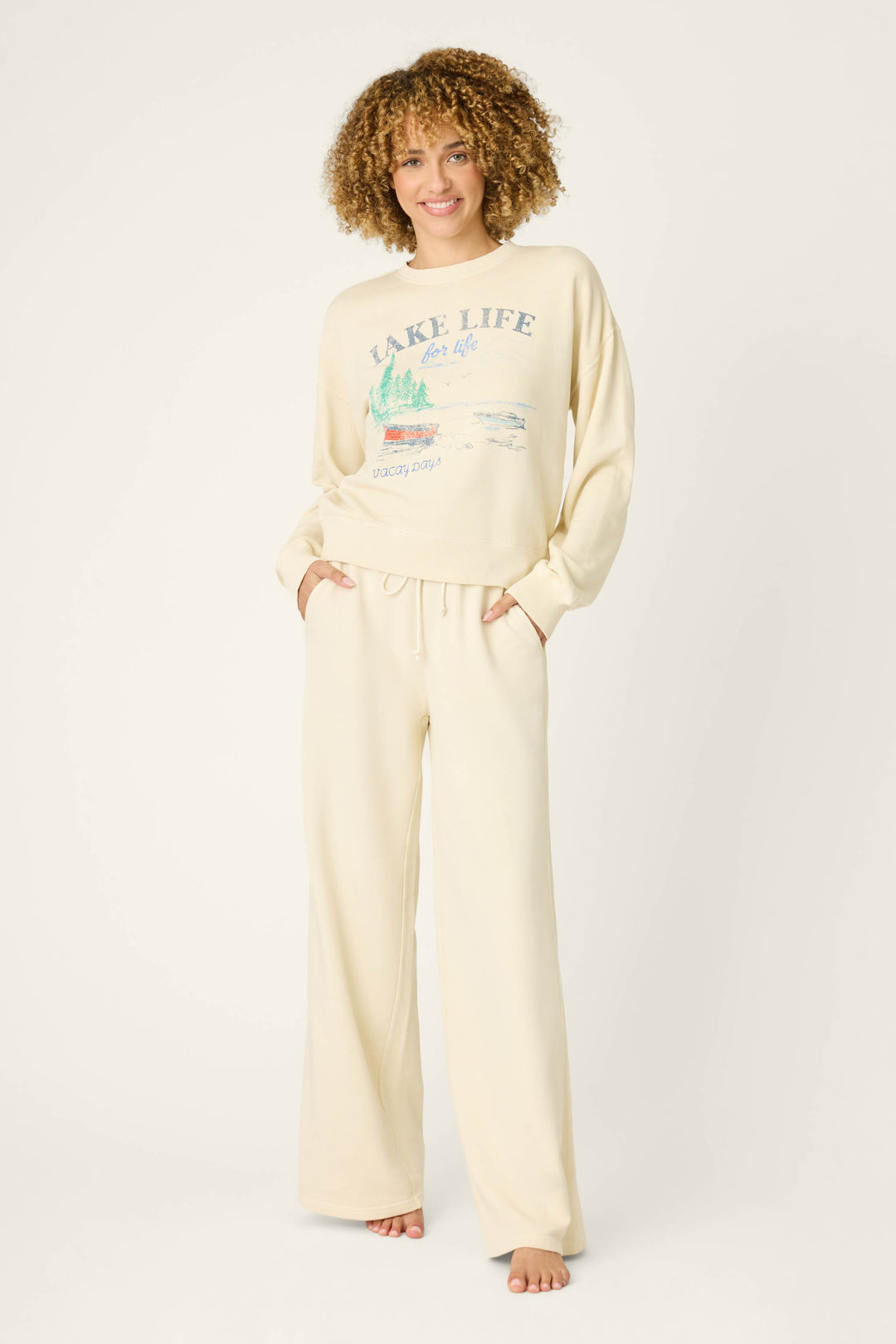 A person with curly hair stands barefoot, hands in pockets, wearing the P.J. Salvage Washed Away Lake Life For Life Long Lounge Set against a plain, light-colored background.