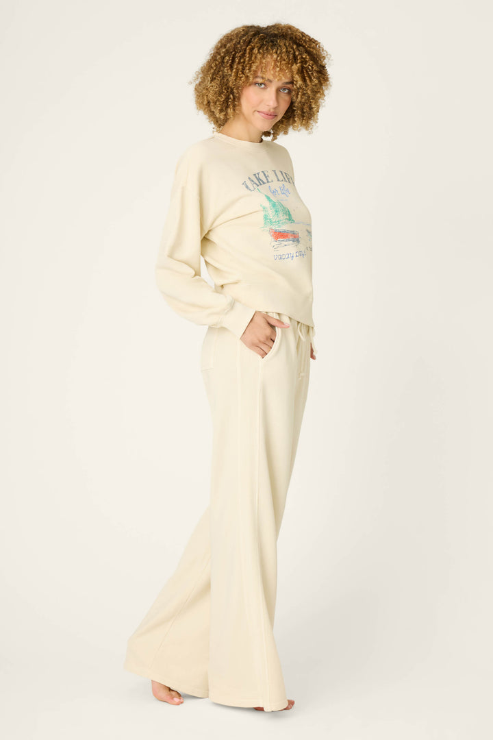 A person with curly hair stands barefoot, hands in pockets, wearing the cream-colored P.J. Salvage Washed Away Lake Life For Life Long Lounge Set—long-sleeve top and wide-leg pants—against a plain light background.