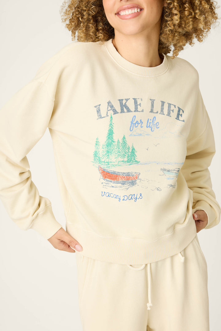 A smiling person poses in the P.J. Salvage Washed Away Lake Life For Life Long Lounge Set, featuring a lakeside graphic with trees and a red boat on cozy light beige French terry—perfect for relaxed vacation days.