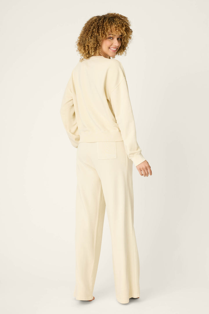 A person with curly hair stands barefoot, smiling over their shoulder in the P.J. Salvage Washed Away Lake Life For Life Long Lounge Set—cream-colored French terry with a long-sleeve top and wide-leg pants—against a plain, light background.