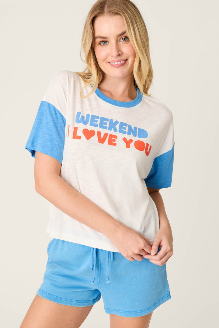 A person wearing the P.J. Salvage Washed Away Weekend I Love You Graphic Tee and blue shorts stands against a plain background, smiling at the camera.