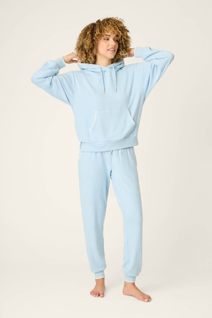 Full-length image of a barefoot model in the Dream Blue hoodie and joggers, hands lifted to the back of the head, relaxed pose.