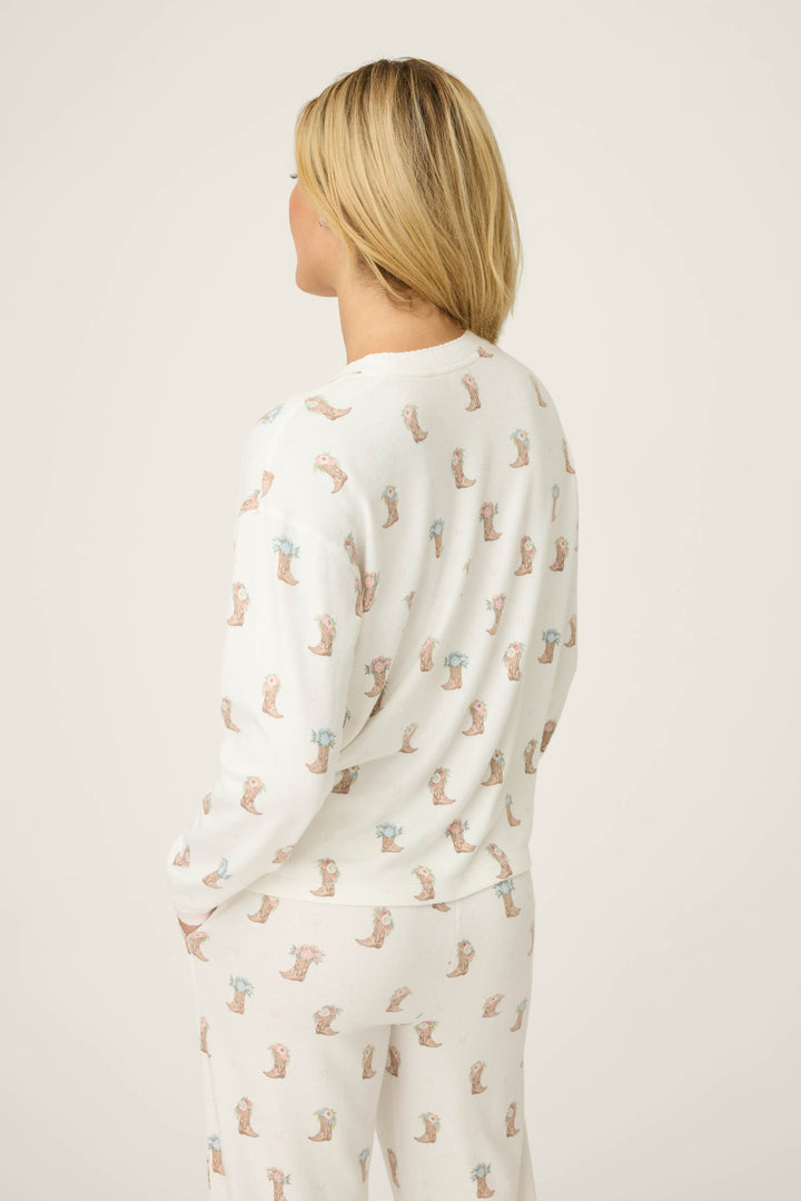 A person with blonde hair is seen from behind wearing the P.J. Salvage Western Blooms Boot Bouquet Long Sleeve Top. The top features a playful pattern, and the background is plain and light-colored.