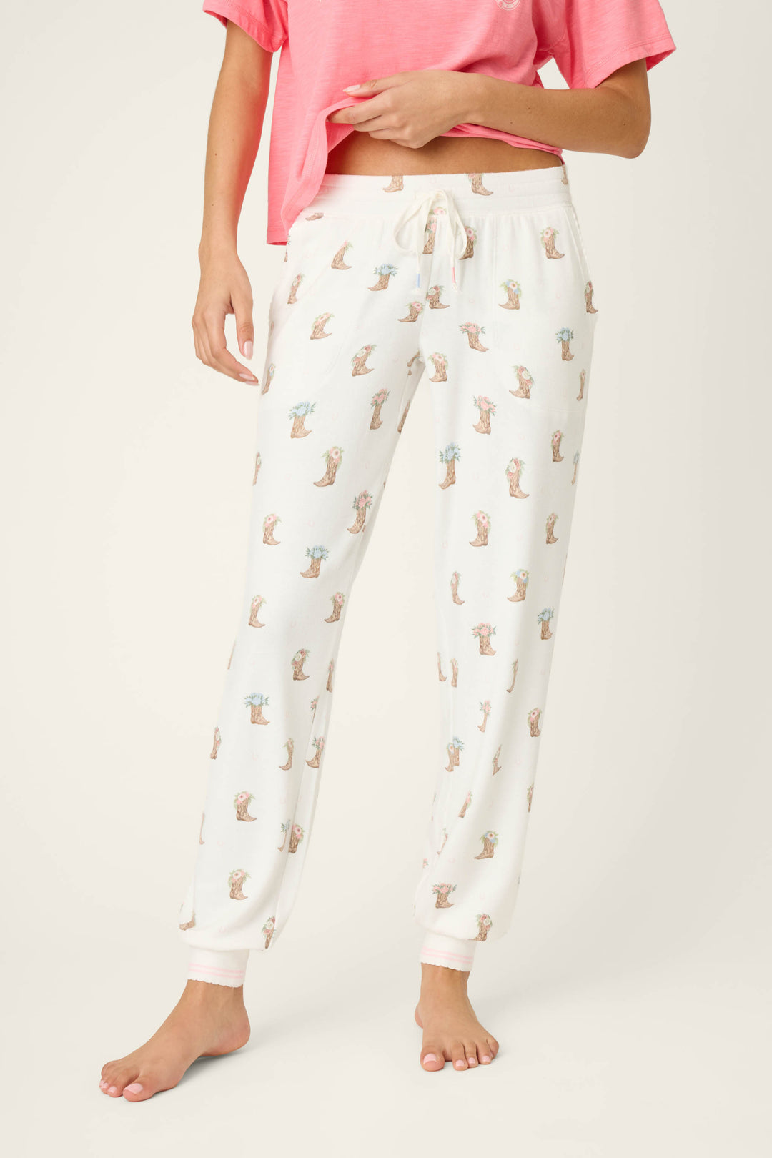 A person wears P.J. Salvage Western Blooms Boot Bouquet Banded Pants, white with a small brown dog print and drawstring waist, paired with a pink short-sleeved top, standing barefoot against a plain light background.