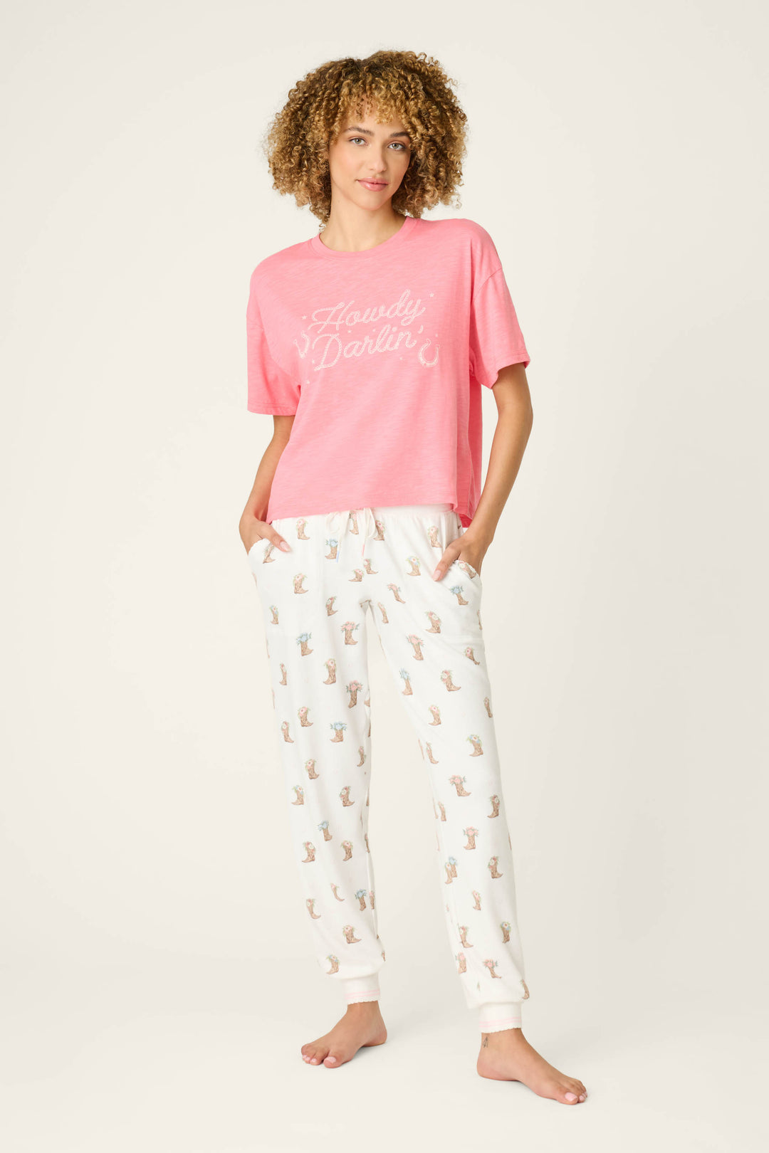 A person stands barefoot against a plain background, wearing a pink “Howdy Darlin” t-shirt and P.J. Salvage’s Western Blooms Boot Bouquet Banded Pants with hands in the pockets.