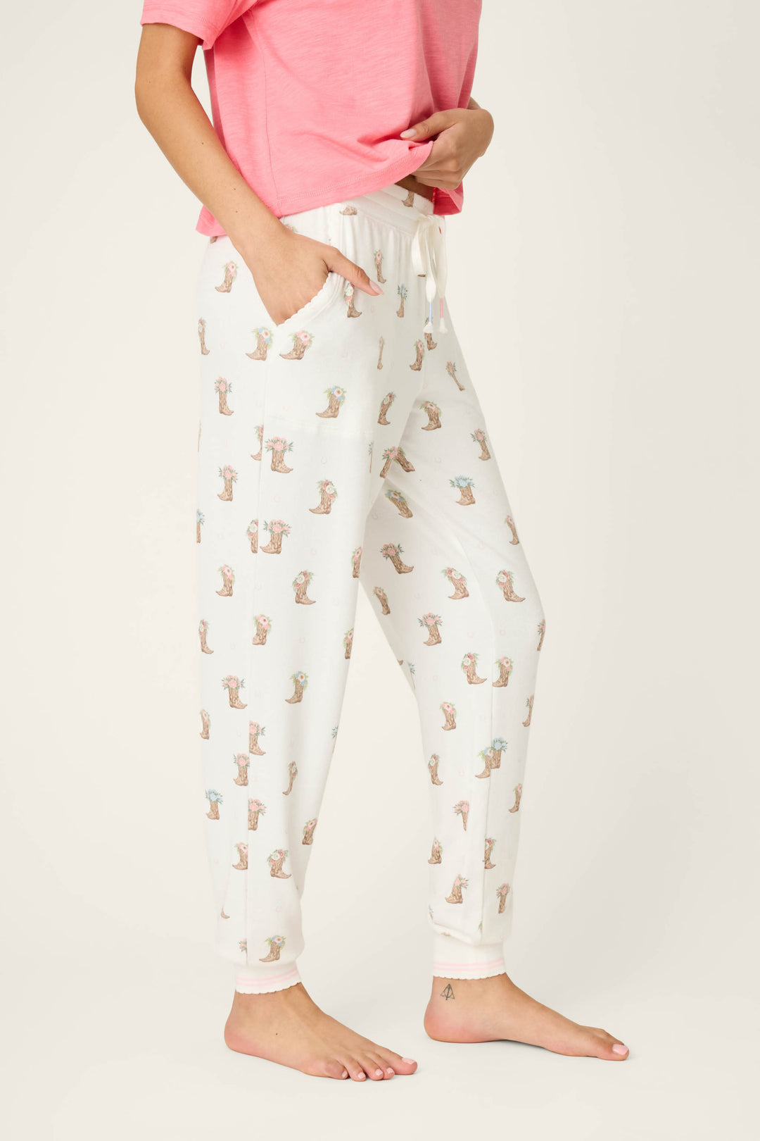 Only the lower half of a person is visible, standing barefoot with one hand in a pocket and the other on their hip, wearing P.J. Salvage Western Blooms Boot Bouquet Banded Pants featuring a small animal print.