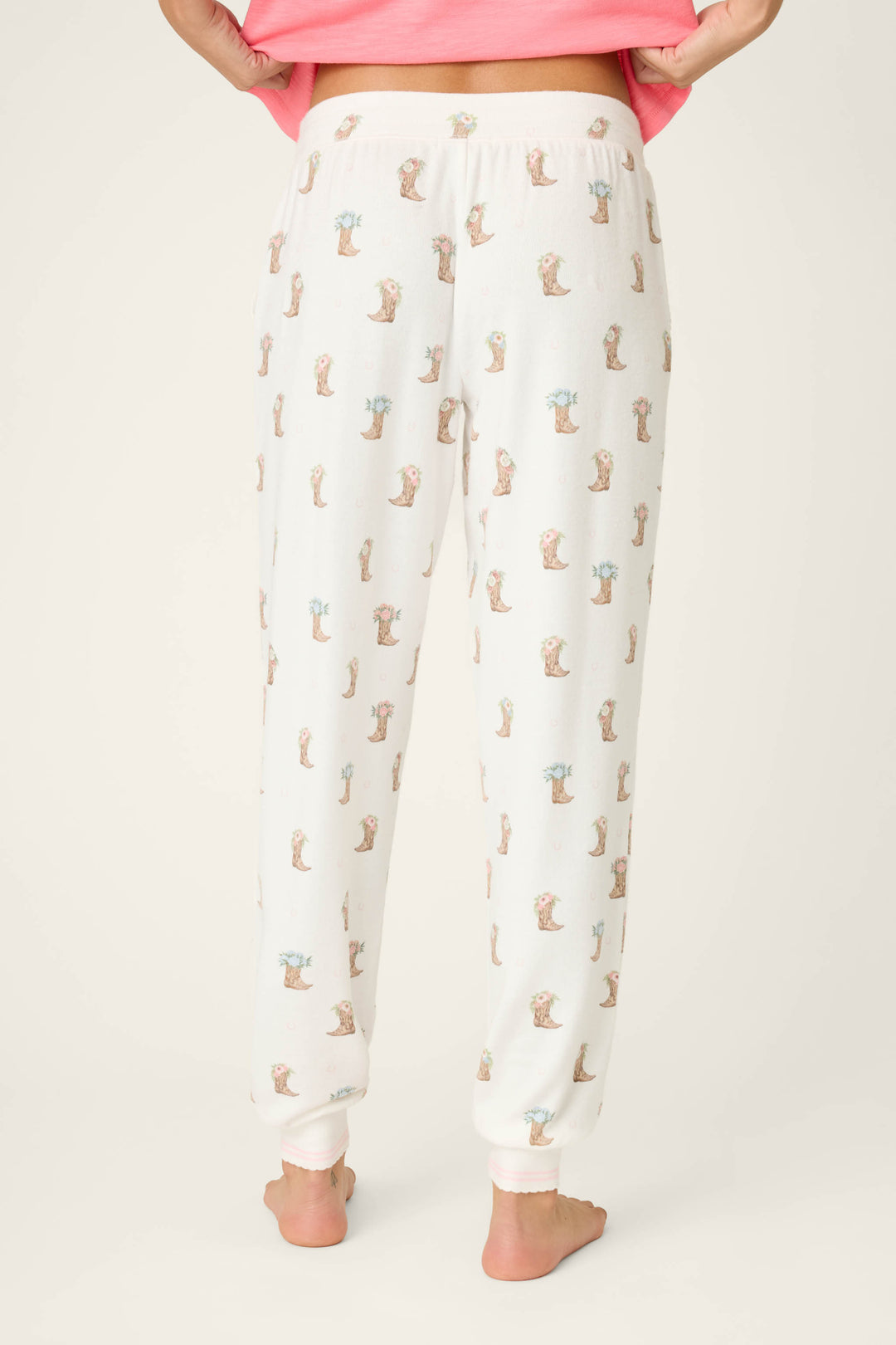A person stands barefoot on a white surface wearing P.J. Salvage Western Blooms Boot Bouquet Banded Pants, featuring a small animal print, and a pink top, shown from the back with hands slightly lifting the hem of the top.