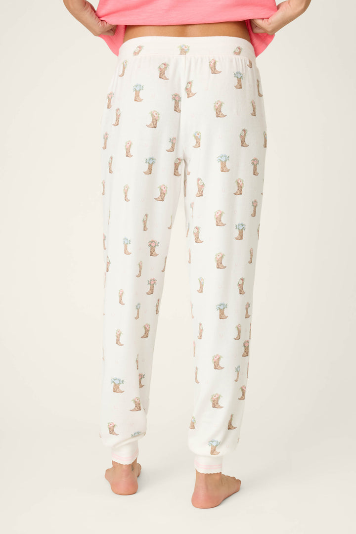 A person stands barefoot on a white surface wearing P.J. Salvage Western Blooms Boot Bouquet Banded Pants, featuring a small animal print, and a pink top, shown from the back with hands slightly lifting the hem of the top.