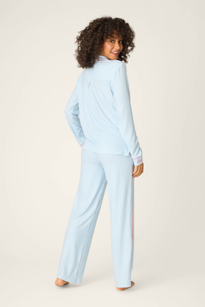 A person with curly hair stands barefoot, facing slightly backward, wearing the P.J. Salvage Western Blooms Blue Stripe Darlin 3-Piece PJ Set featuring light blue and white stripes and pink accents on the cuffs and seams.